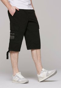 Černé cargo šortky s kapsami, texturovan�ý materiál a nastavitelné lemy. Spárováno s bílými a šedými sportovními botami se značkováním.