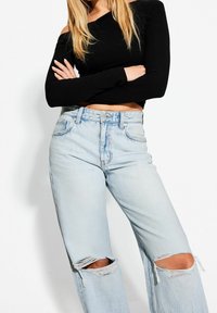 Musta olkapäät paljastava pitkähiha toppi, yhdistetty vaaleansinisiin revittyihin leveälahkeisiin farkkuihin, joissa on rispaantuneet reunat polvaukkojen ympärillä.