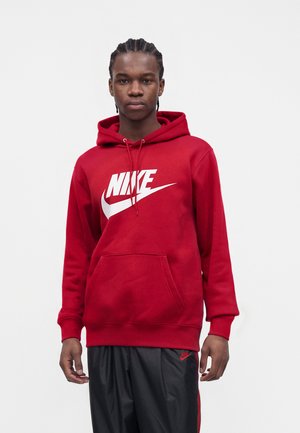 Junger Mann mit geflochtenem Haar, der einen roten Nike-Hoodie und schwarze Nike-Sweatpants trägt, steht vor einem schlichten Hintergrund.