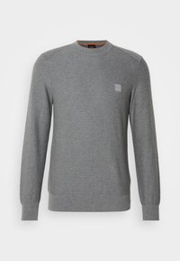 Pull tricoté gris à manches longues avec col rond et petite pièce décorative sur la poitrine gauche, présenté sur un fond blanc.