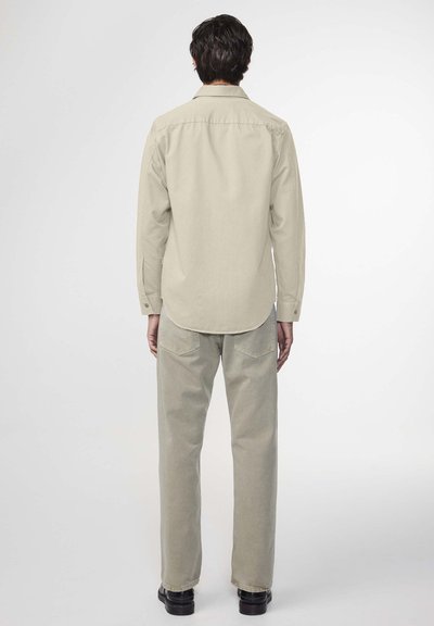 Beige Langarmhemd mit abgerundetem Saum, kombiniert mit hellbeigen geraden Hosen. Die Materialien wirken weich und haben minimalistische Designdetails.