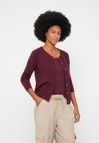 Cárdigan burdeos con un escote redondo y seis botones, fabricado en un tejido suave. Combinado con una blusa de canalé y pantalones cargo beige.