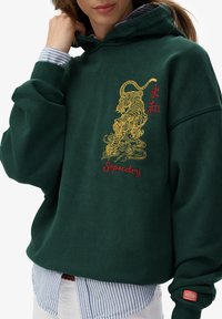 Vrouw draagt een donkergroene hoodie met een gele tijgerborduring en rode "Superdry" tekst, gedragen over een blauw-wit gestreept overhemd.