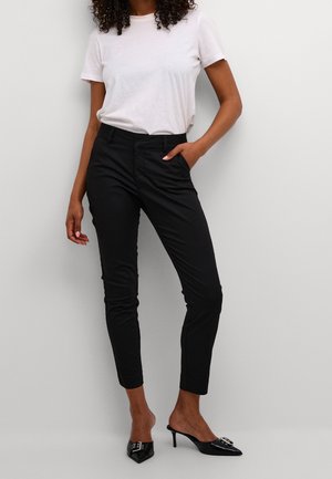 Vrouw die een wit T-shirt met korte mouwen draagt, strakke zwarte broek tot op de enkel en zwarte slip-on schoenen met hoge hakken en puntige neuzen.