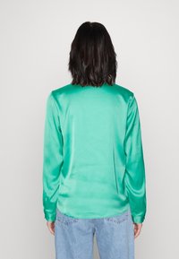 Top lungo a maniche e satin verde con texture liscia, collo rotondo e orlo leggermente curvo, abbinato a jeans in denim blu chiaro.