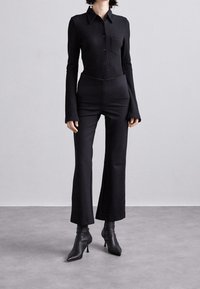 Schwarzes, geripptes Langarmhemd mit Knopfleiste und Tasche, kombiniert mit hochtaillierten, weit geschnittenen schwarzen Hosen und schwarzen Ankle Boots mit spitzem Kopf.