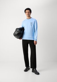 Light blue sweatshirt with black trim and a front graphic, paired with black trousers and black lace-up shoes. Holding a black bag.