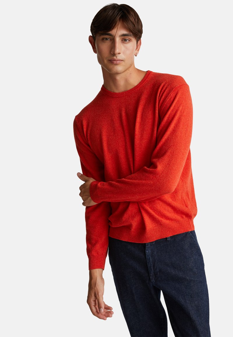 Młody mężczyzna z krótkimi ciemnymi włosami, ubrany w jasnoczerwony sweter i ciemnoniebieskie dżinsy, pozujący z jednym ramieniem skrzyżowanym na ciele.