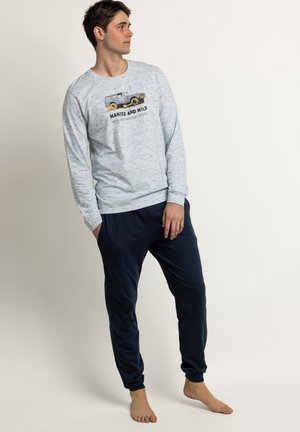 Joven descalzo con sudadera gris clara con gráfico de coche y pantalones azul oscuro, mirando hacia su izquierda sobre fondo liso.