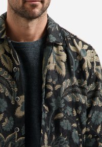 Jasje met bloemenpatroon in donkere stof, met tinten blauw en goud. Het heeft een kraag en knoopsluiting, gelaagd over een donkere trui.