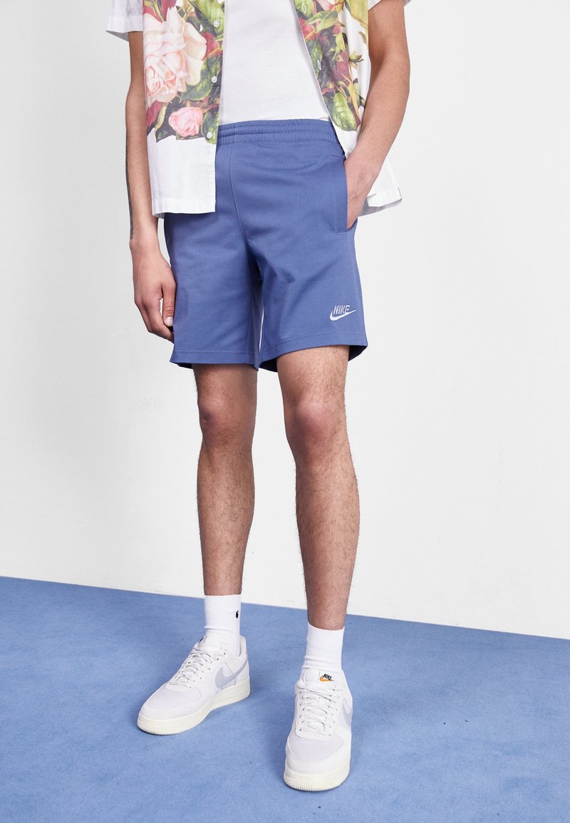 Personne portant un short Nike bleu, des chaussettes blanches, des baskets Nike blanches, un débardeur blanc et une chemise fleurie ouverte, debout sur un sol bleu.