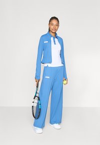 Blå sportsoutfit bestående af en tætsiddende jakke og løse bukser, parret med en hvid polo-shirt, mens der holdes en tennisracket og bold.