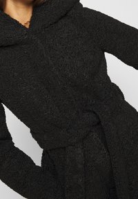 Svart, texturerad huva kappa gjord av mjukt tyg med ett knytbälte vid midjan. Har en framträdande huva och ett mysigt, plyschigt utseende.