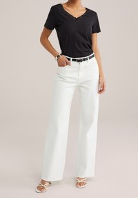 Black V-neck t-shirt with lace detailing, paired with high-waisted white wide-leg jeans. White strappy sandals and a black belt accent the outfit.