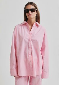 Camicia button-up a maniche lunghe di colore rosa chiaro con vestibilità rilassata, caratterizzata da colletto classico e maniche lunghe. Tessuto di cotone strutturato.