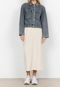 Persona che indossa una giacca di jeans grigia corta, una gonna midi beige con tasche laterali e sneakers bianche, in piedi su un pavimento semplice contro una parete bianca.