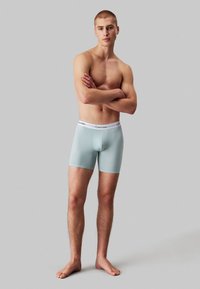 Lichtblauwe stretch boxers met een witte Calvin Klein tailleband, met een strakke pasvorm en een gladde textuur. Model poseert met gekruiste armen, blote voeten.