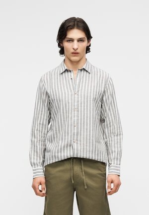 Ung mand iført en grå og hvid lodretstribet skjorte med knapper og olivengrønne bukser med snøre, stående mod en ensfarvet baggrund.