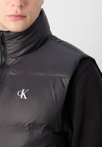 Gilet imbottito nero con colletto alto, caratterizzato da una finitura lucida e un piccolo logo bianco 'cK' sul petto, indossato sopra una maglietta nera a maniche lunghe.