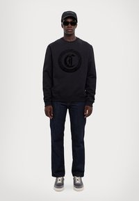 Felpa nera con logo in rilievo, jeans blu scuro, sneakers grigie e cappellino nero. L'outfit presenta un design informale e un tessuto testurizzato.