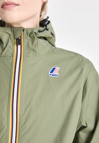 Giacca leggera verde K-Way con colletto alto, dotata di zip centrale decorata con righe nere, bianche e gialle, e una toppa con logo.