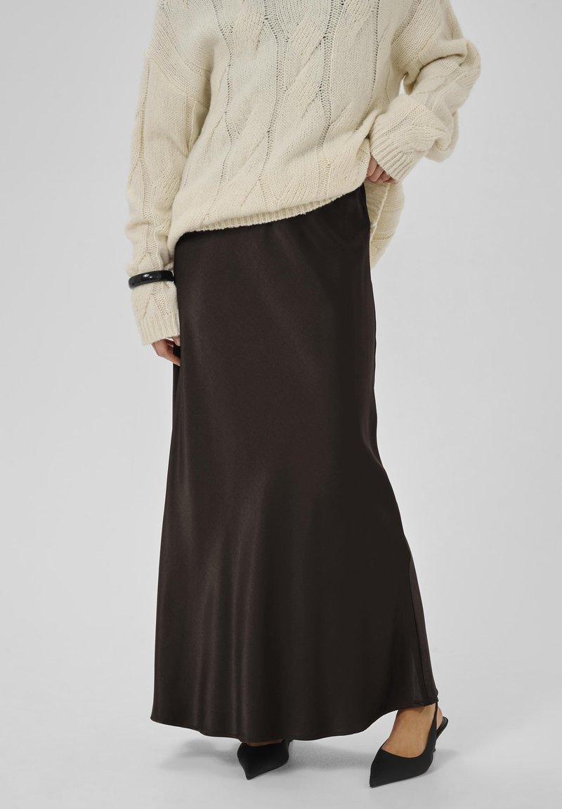 Lang, mørkebrun satin-nederdel med en glat tekstur og en sidedel, samt en oversized cremet kabelstriktrøje og sorte slingback-sandaler.