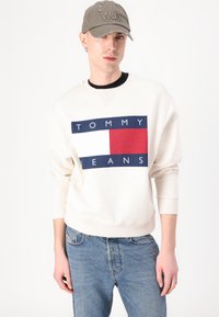 Krémová mikina s modrým a červeným logom "TOMMY JEANS", kombinovaná so svetlomodrými džínsami. Model nosí sivú čiapku s textom "Won".
