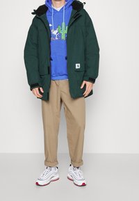 Persoon draagt een donkergroene capuchonjas, een blauwe hoodie met een cactusprint, beige broek en witte sneakers, staand tegen een effen achtergrond.