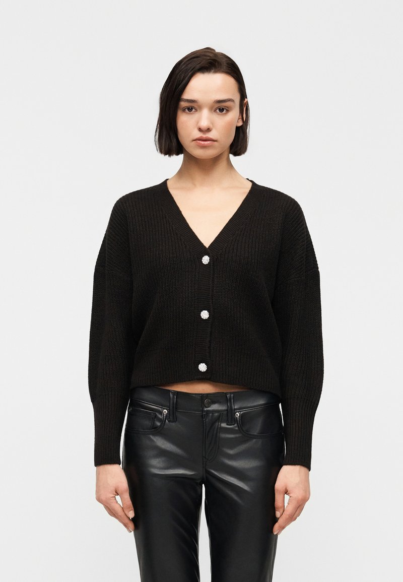 Γυναίκα με κοντά σκούρα μαλλιά φοράει μαύρο κοντό κουμπωτό ζακέτα και μαύρο δερμάτινο παντελόνι, στέκεται μπροστά σε απλό φωτεινό φόντο.