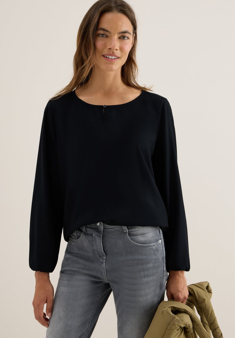 Zwarte langemouwen top met een ronde halslijn en een klein sleutelgatdetail, gecombineerd met grijze aansluitende jeans. Soepele textuur, comfortabele pasvorm.