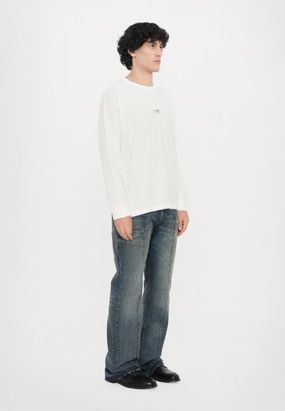 Vit långärmad t-shirt med en liten grafik vid bröstet, kombinerad med bleka blå utsvängda jeans och svarta skor. Enkelt bakgrund.