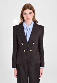 Blazer marrone doppiopetto con bottoni dorati, rever a scollatura, e due tasche laterali. Indossato sopra una camicia azzurro chiaro a righe.