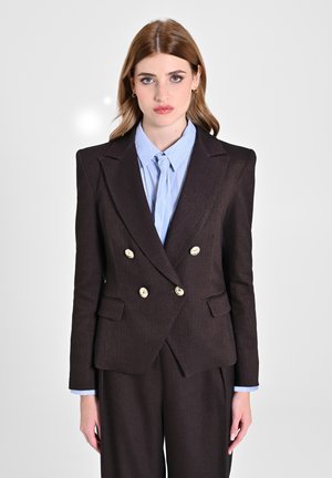 Blazer marrone doppiopetto con bottoni dorati, rever a scollatura, e due tasche laterali. Indossato sopra una camicia azzurro chiaro a righe.