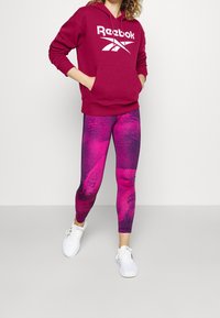 Maroon Reebok-hoodie med en ficka fram och vit logotyp, i kombination med rosa leggings med mönstrad textur, och vita träningsskor.