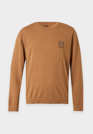 Junger Mann steht, trägt ein braunes Langarmshirt, ausgewaschene blaue Jeans und grüne Sneakers mit orangen Schnürsenkeln vor weißem Hintergrund.