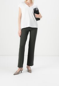 Bílá tričko s V výstřihem, měkká látka, v kombinaci s tmavě zelenými kalhotami rovného střihu. Modelka drží černou kabelku; šedé sandály na podpatku dokončují outfit.