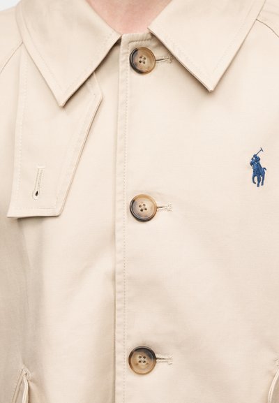 Μπεζ παλτό τύπου trench με κλασικό γιακά, δύο κουμπώματα με κλωστές σε αντίθετο χρώμα και κεντημένο λογότυπο σε ναυτικό μπλε.