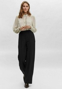 Camicia bianca con pois neri e colletto arricciato, abbinata a pantaloni a vita alta neri a gamba larga. Tessuto morbido, vestibilità sartoriale.