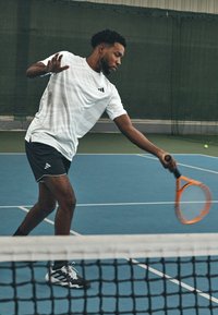 Valge sportline särg, millel on peened mustrid, mustad lühikesed püksid ja mustad tenniseciped. Hoides käes oranži tennisereketit, on ta valmis palli lööma.