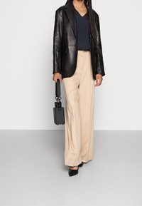 Blazer en cuir noir porté sur un chemisier bleu marine, associé à un pantalon large beige et des escarpins noirs à bout pointu. Un petit sac à main noir est tenu.