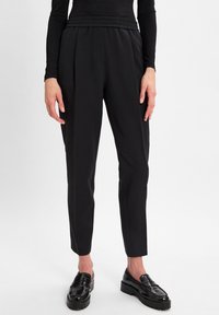 Persoon in zwarte taps toelopende broek, zwart lang mouwtop en glanzende zwarte loafers die op een witte vloer staat.