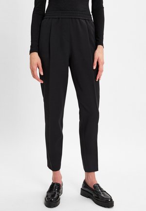 Persoon in zwarte taps toelopende broek, zwart lang mouwtop en glanzende zwarte loafers die op een witte vloer staat.