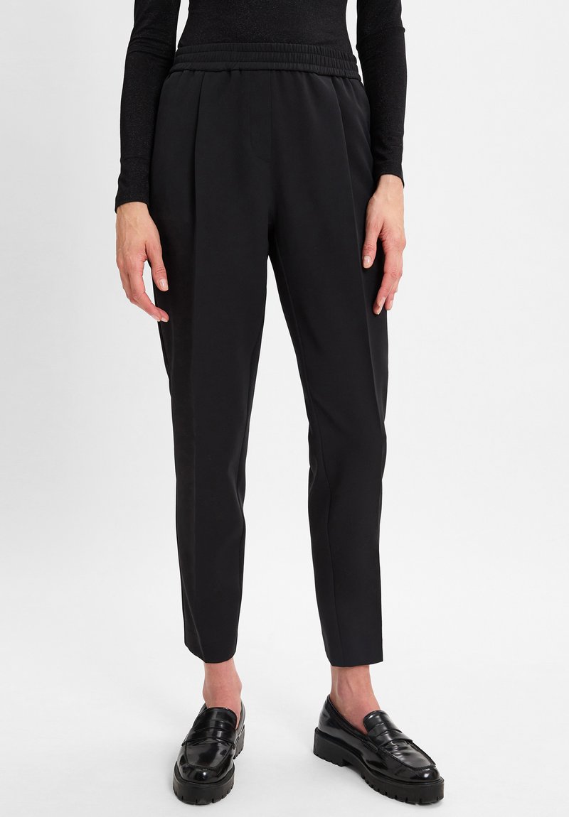 Persoon in zwarte taps toelopende broek, zwart lang mouwtop en glanzende zwarte loafers die op een witte vloer staat.