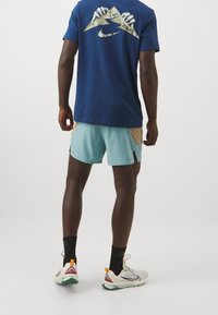 Camiseta azul de Nike con gráfico de montaña, pantalones cortos azul claro con acentos beige, calcetas negras y zapatillas deportivas multicolores.