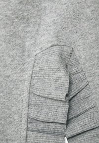 Grauer Strickpullover mit gerippter Textur und ausgestellten Rüschendetails an den Ärmeln, die die geschichtete Bewegung des Stoffs betonen.