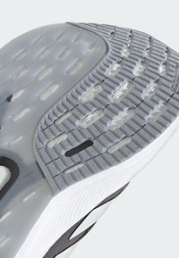Sola della scarpa con una texture in gomma grigia, dotata di una sezione traslucida e schemi circolari per la trazione. Tallone arrotondato e scanalature strutturali presenti.