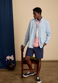 Lichtblauw overhemd met knopen over een roze graphic t-shirt, marineblauwe shorts en marineblauwe sneakers. Model leunt tegen een stoel met een bloemenvaas in de buurt.