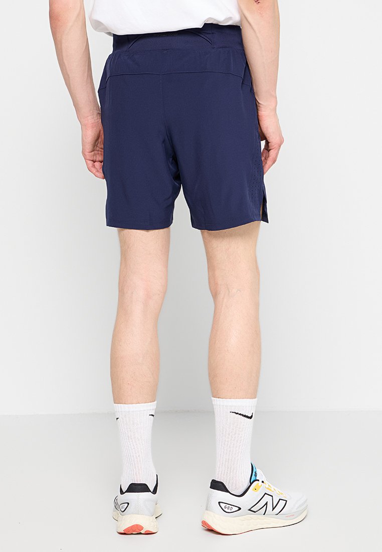 Persoon in marineblauwe sportshorts, witte crew-sokken met zwart logo, en witte hardloopschoenen met kleurrijke accenten, staande tegen een witte achtergrond.