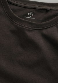 Tumšbrūnas t-krekls ar apaļu kakla izgriezumu. Izgatavots no mīksta auduma; satur birku ar uzrakstiem "THE SET M" un "MADE IN BANGLADESH."