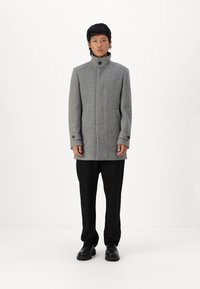 Cappotto grigio in misto lana con colletto alto, chiusura frontale con bottoni e tasche laterali. Indossato sopra pantaloni e scarpe neri. Design semplice e fitted.
